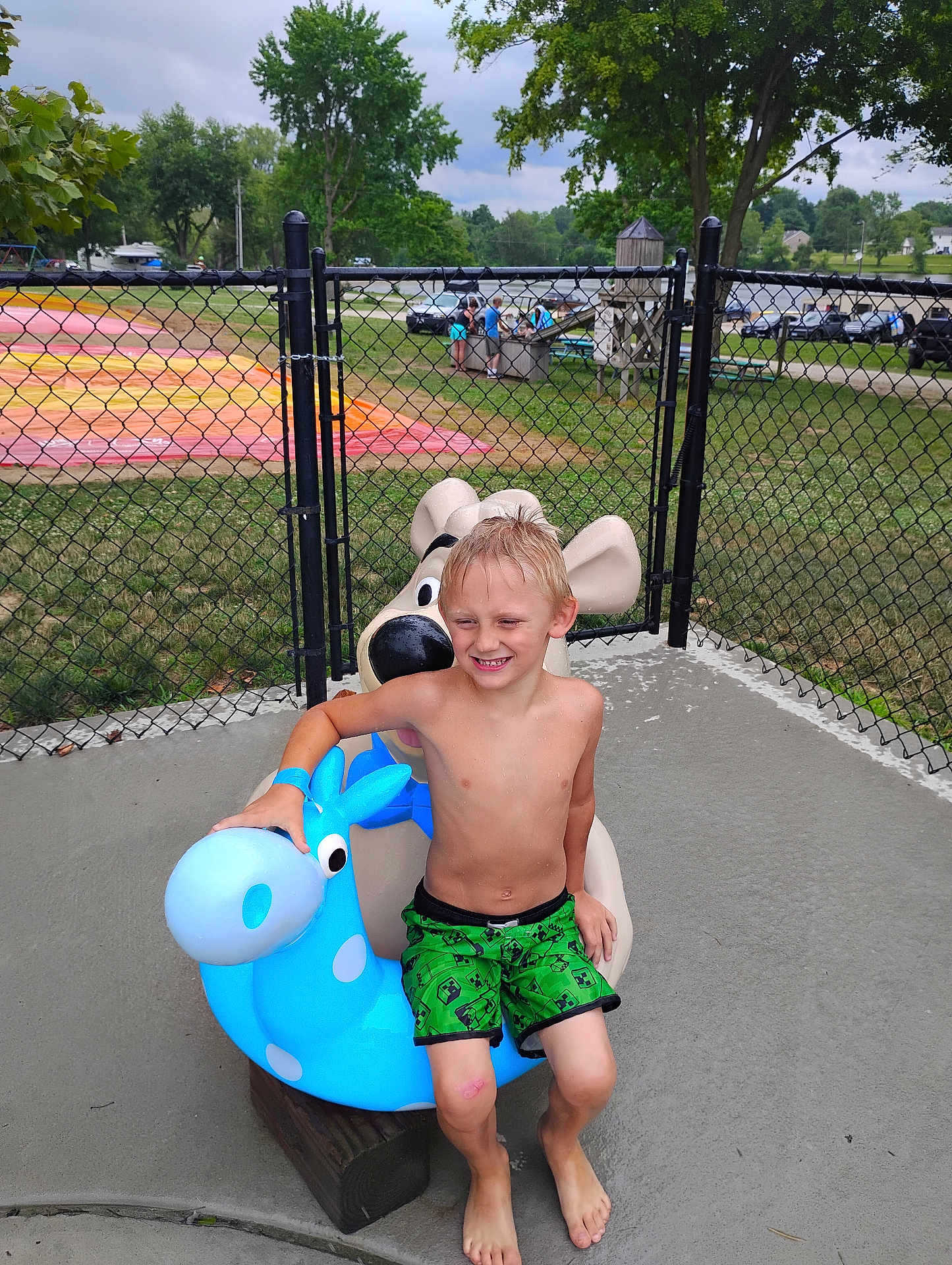 Jacob is registered to the contest to win money with this photo: child, boy, smile, playground, play_equipment, outdoor, summer, wet_hair, green_shorts, barefoot, fence, trees, grass, concrete, toy, fun, happy, park, daytime, casual