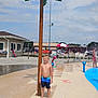 child, boy, water_sprinkler, splash_pad, swim_trunks, barefoot, outdoor, summer, pool_area, umbrella, chairs, water_droplets, palm_tree, playground, sunny, recreation, fun, leisure, family, daytime