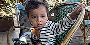 Othman a rejoint le concours — aidez-le/la à gagner de superbes lots ! child, toddler, striped_sweater, teddy_bear_patch, green_wicker_chair, outdoor, cafe, stroller, curly_hair, wooden_floor, table, plants, daylight, person, sitting, casual_clothing, cute, young_child, holding, expression