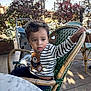 child, toddler, striped_sweater, teddy_bear_patch, green_wicker_chair, outdoor, cafe, stroller, curly_hair, wooden_floor, table, plants, daylight, person, sitting, casual_clothing, cute, young_child, holding, expression