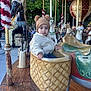 Othman participe au concours pour gagner de l'argent avec cette photo : toddler, child, carousel, amusement_ride, horse, hat, jacket, outdoor, wooden_floor, seat, face, expression, festive, decoration, tree, daylight, smile, park, playful, clothing