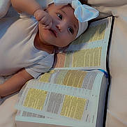 Genesis is registered to the contest to win money with this photo: baby, infant, headband, white_bow, book, open_book, text, bed, blanket, soft, cute, child, portrait, indoors, resting, hand, face, eyes, clothing, peaceful