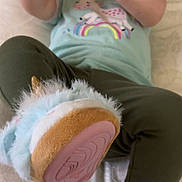 Ella joined the competition — help win amazing prizes! baby, child, toy, unicorn_slippers, green_shirt, pants, bed, blanket, hand, footwear, indoor, person, infant, cute, plush, sleepwear, reaching, soft_toys, colorful, lying_down