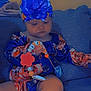 baby, beads, blue_clothing, child, comfort, couch, cute, flower, focus, headband, homedecor, indoor, infant, orange_clothing, person, playful, portrait, sitting, soft_lighting, toy