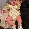 baby, infant, headband, bow, romper, yellow, boots, flower_pattern, blanket, pillow, plaid, sitting, cute, indoors, child, person, clothing, footwear, expression, portrait