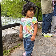 Saanvhi is registered to the contest to win money with this photo: toddler, child, curly_hair, tie_dye_shirt, pink_sandals, jeans, outdoor, river, stone_path, greenery, trees, railing, people, casual_clothing, nature, daylight, side_view, curious_expression, family, walking_area