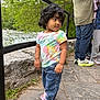 toddler, child, curly_hair, tie_dye_shirt, pink_sandals, jeans, outdoor, river, stone_path, greenery, trees, railing, people, casual_clothing, nature, daylight, side_view, curious_expression, family, walking_area