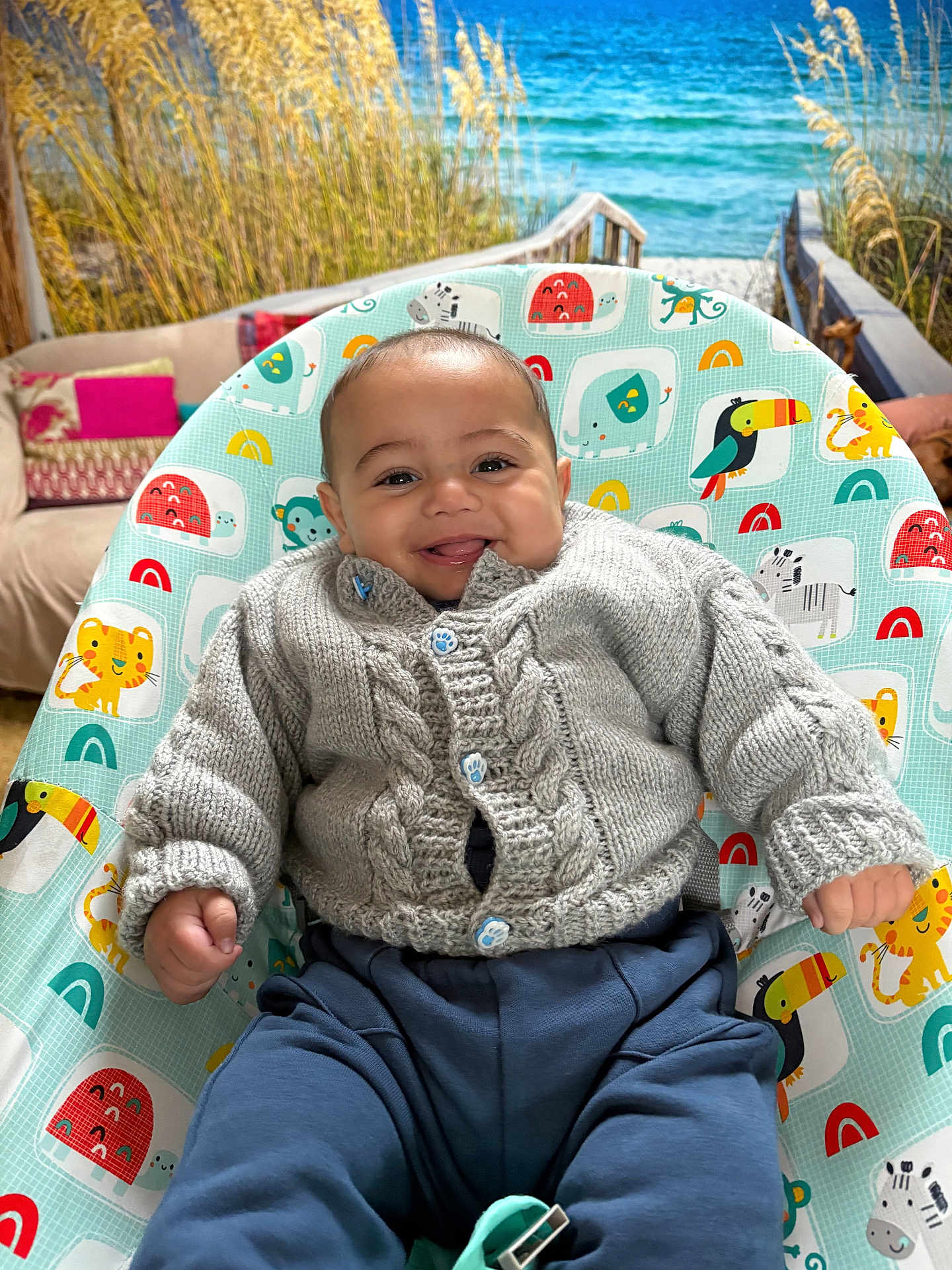 Flynn joined the competition — help win amazing prizes! baby, smiling, knitted_sweater, blue_pants, baby_seat, animal_pattern, indoor, couch, pillows, beach_mural, ocean, grass, happy, child, portrait, cozy, cute, infant, seat, playful