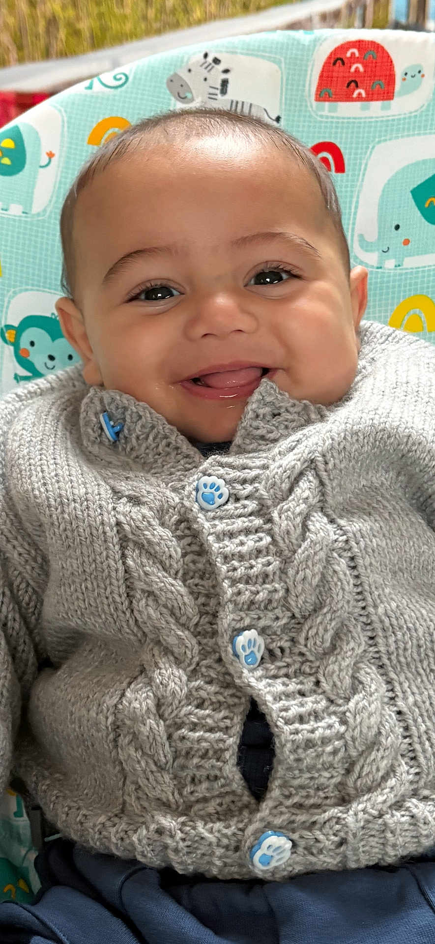 Flynn is registered to the contest to win money with this photo: baby, smiling, face, knitted_sweater, button, paw_print, high_chair, animal_pattern, cute, child, indoor, portrait, happy, clothing, infant, closeup, seated, warm, cozy, playful