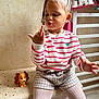child, braided_hair, striped_shirt, shorts, tights, toy, dog_toy, bench, polka_dots, minnie_mouse_bag, indoor, wall, floor, playful, cute, person, sitting, expression, earrings, furniture