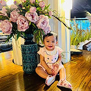 Sareen is registered to the contest to win money with this photo: baby, child, smiling, flowers, vase, pink, dress, sandals, wooden_table, indoor, decor, green_leaves, happy, sitting, person, cute, ornate, plant, lighting, bracelet