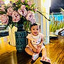 baby, child, smiling, flowers, vase, pink, dress, sandals, wooden_table, indoor, decor, green_leaves, happy, sitting, person, cute, ornate, plant, lighting, bracelet