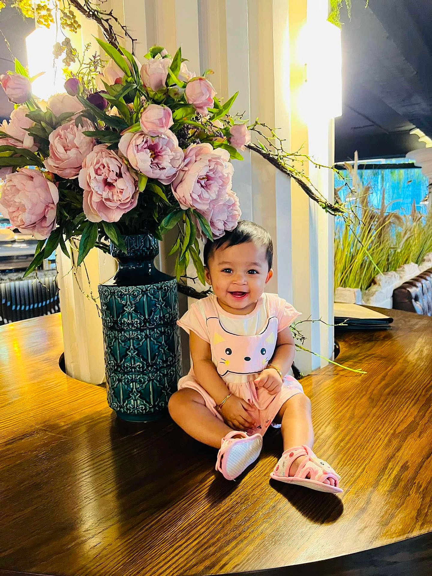 Sareen is registered to the contest to win money with this photo: baby, child, smiling, flowers, vase, pink, dress, sandals, wooden_table, indoor, decor, green_leaves, happy, sitting, person, cute, ornate, plant, lighting, bracelet