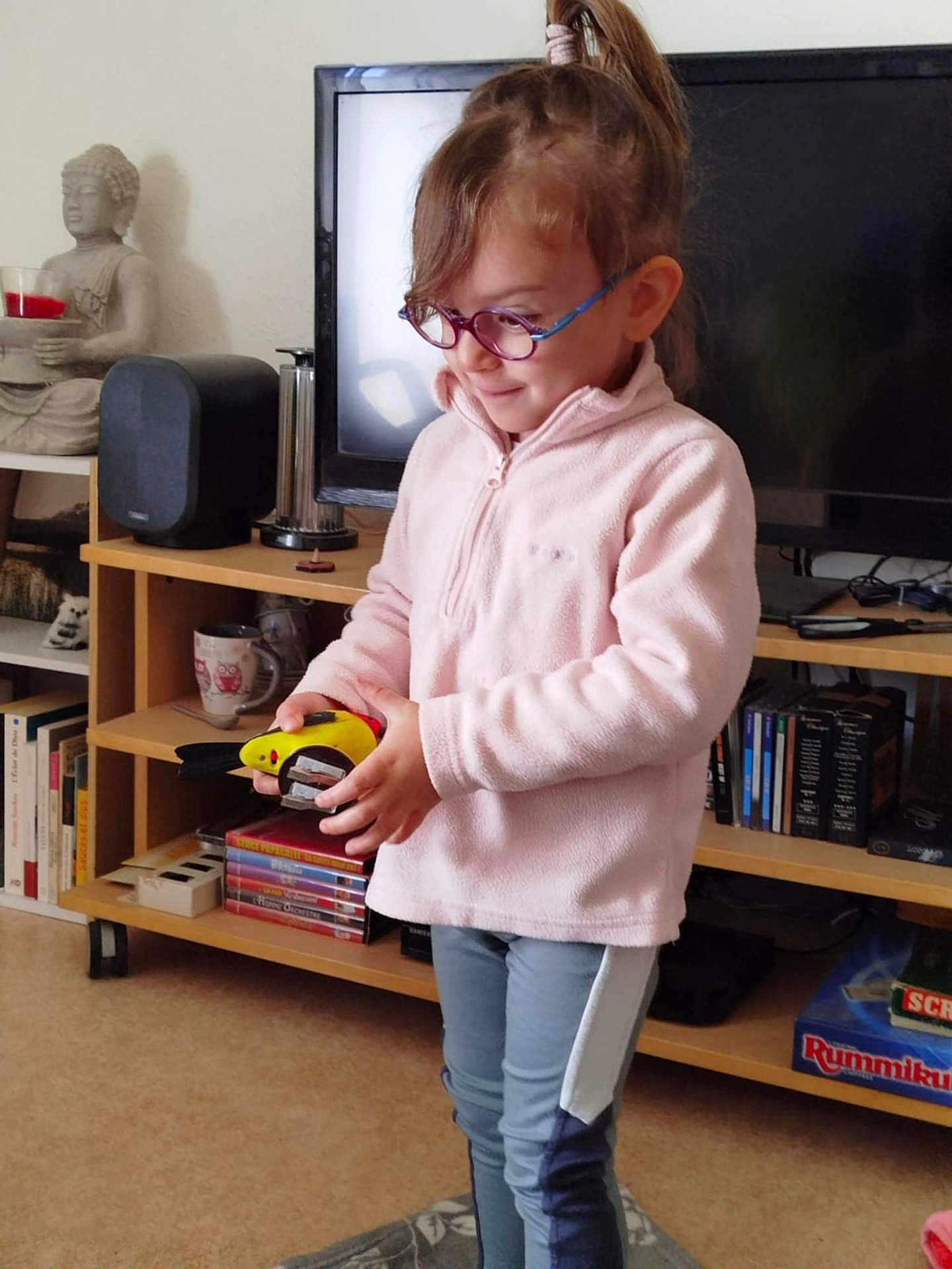 Kena a rejoint le concours — aidez-le/la à gagner de superbes lots ! child, glasses, toy, living_room, bookshelf, television, statue, buddha, sweater, pants, curly_hair, carpet, shelf, cup, indoor, person, furniture, curiosity, casual_clothing, play