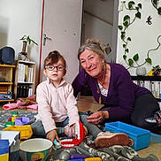 Kena participe au concours pour gagner de l'argent avec cette photo : child, adult, woman, glasses, toys, indoor, blanket, smile, bookshelf, plant, floor, playing, person, casual_clothing, room, door, carpet, headwear, footwear, toy_blocks