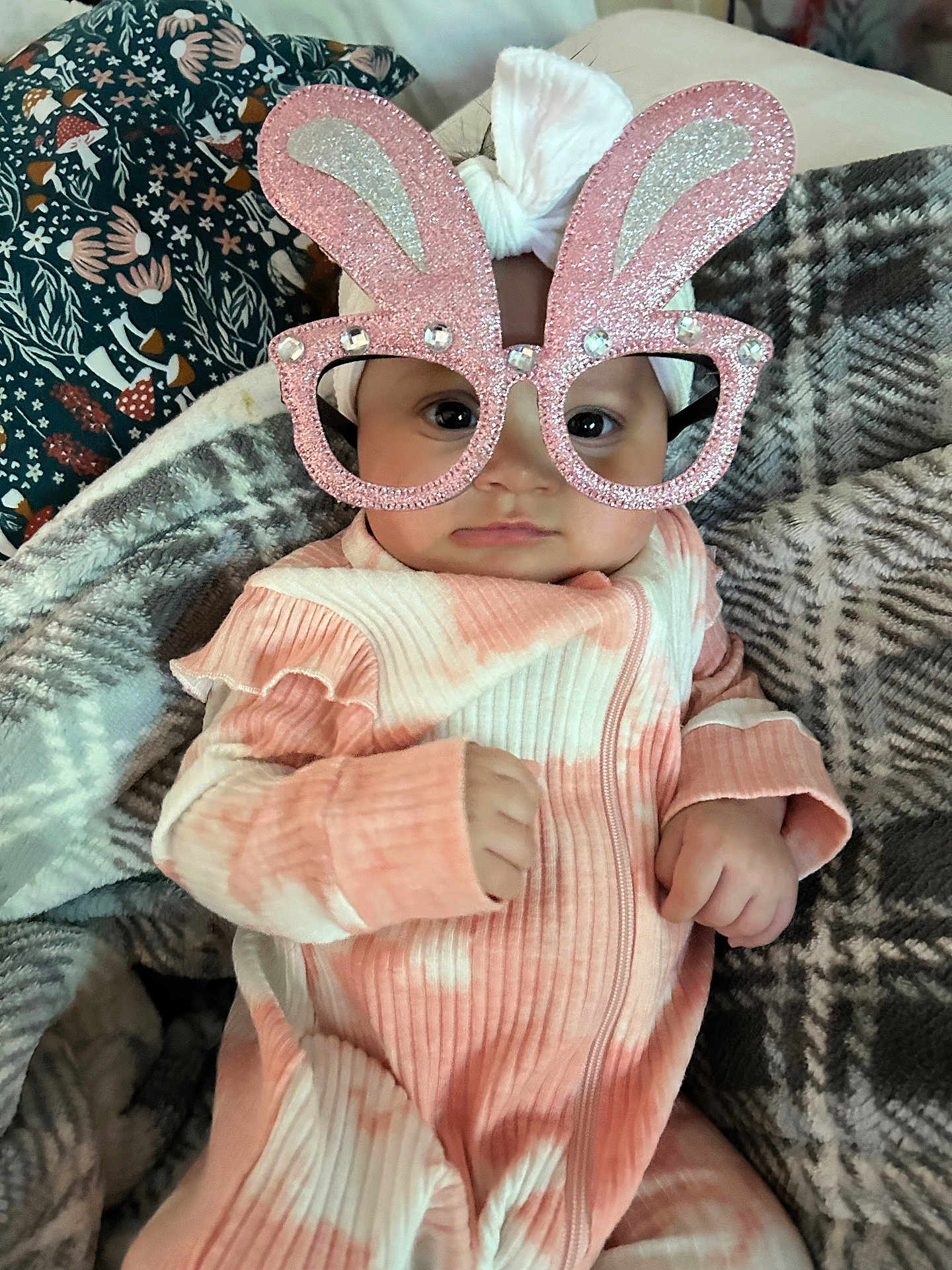 Gia is registered to the contest to win money with this photo: baby, infant, glasses, bunny_ears, headband, pink_clothing, blanket, patterned_blanket, cute, face, hands, indoor, cozy, child, portrait, fashion_accessory, soft_texture, baby_girl, expression, closeup