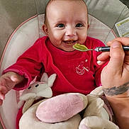 Inaya a rejoint le concours — aidez-le/la à gagner de superbes lots ! baby, infant, feeding, spoon, green_food, red_clothing, smiling, plush_toys, soft_toys, baby_seat, cushion, happy, cute, hand, tattoo, close_up, indoor, person, child, portrait