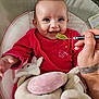 baby, infant, feeding, spoon, green_food, red_clothing, smiling, plush_toys, soft_toys, baby_seat, cushion, happy, cute, hand, tattoo, close_up, indoor, person, child, portrait