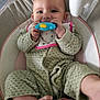 baby, infant, toy, chewing, green_outfit, bib, baby_seat, cushion, smiling, happy, cute, barefoot, indoor, floor, carpet, child, person, playful, closeup, portrait