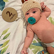 Kyzer is registered to the contest to win money with this photo: baby, infant, pacifier, knitted_hat, blanket, wooden_sign, 2_months, cute, lying_down, raised_fist, soft_texture, patterned_blanket, milestone, adorable, child, headwear, celebration, indoor, portrait, young