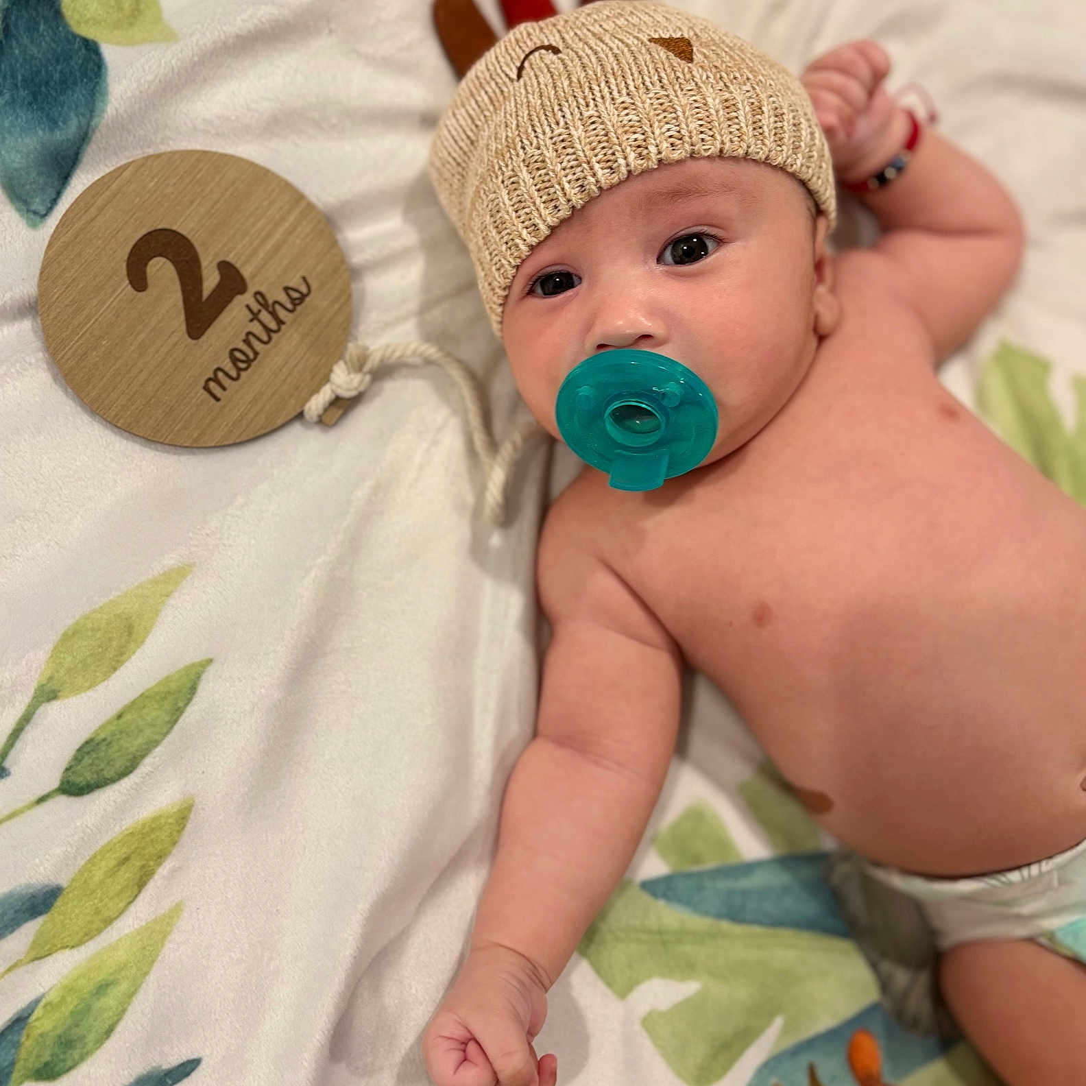 Kyzer is registered to the contest to win money with this photo: 2_months, adorable, baby, blanket, celebration, child, cute, headwear, indoor, infant, knitted_hat, lying_down, milestone, pacifier, patterned_blanket, portrait, raised_fist, soft_texture, wooden_sign, young