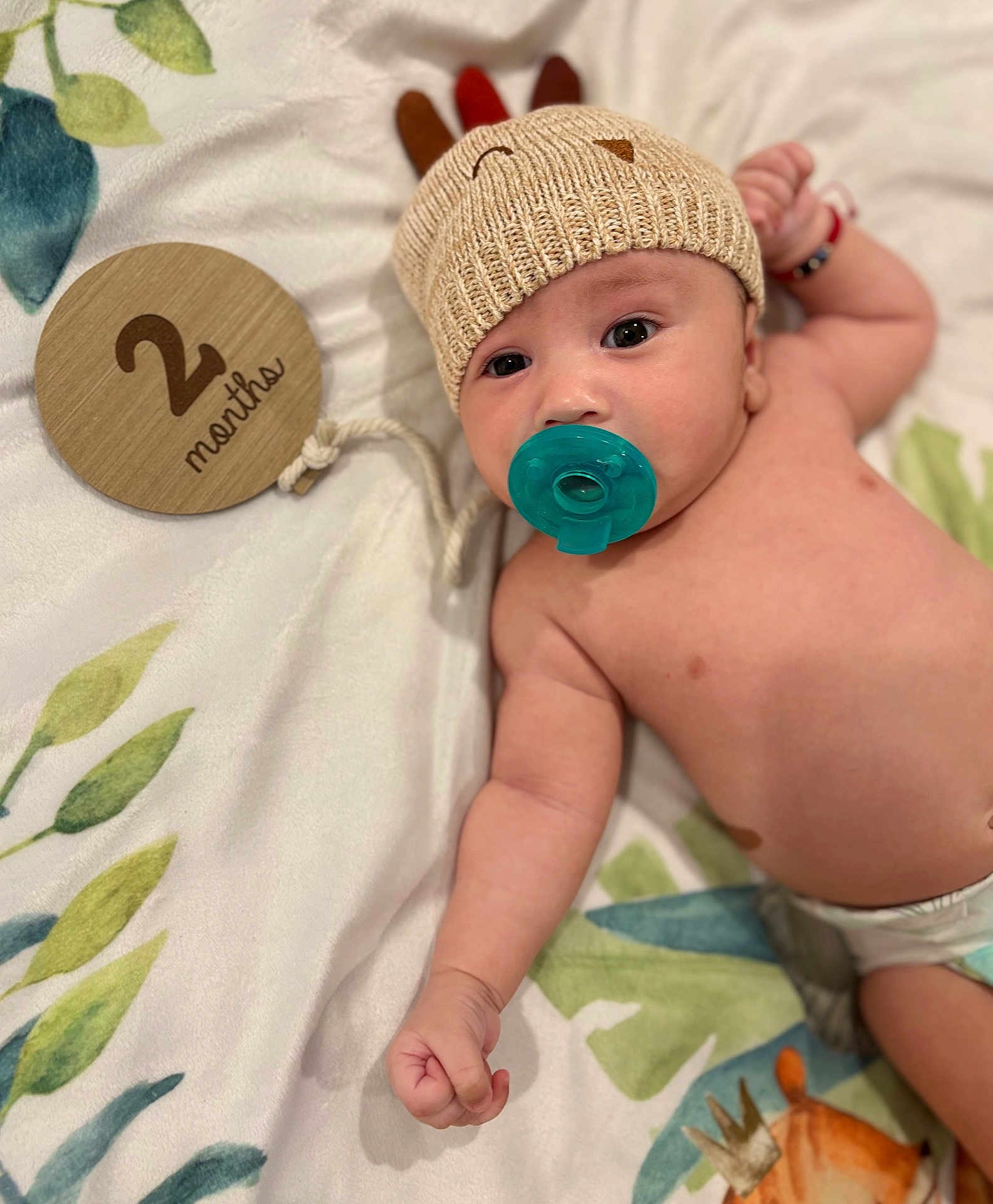 Kyzer is registered to the contest to win money with this photo: baby, infant, pacifier, knitted_hat, blanket, wooden_sign, 2_months, cute, lying_down, raised_fist, soft_texture, patterned_blanket, milestone, adorable, child, headwear, celebration, indoor, portrait, young