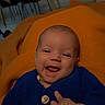 baby, blue_sweater, button, chair, clothing, cozy, cute, dining_area, face, furniture, hand, happy, indoor, infant, orange_blanket, person, portrait, skin, smiling, table