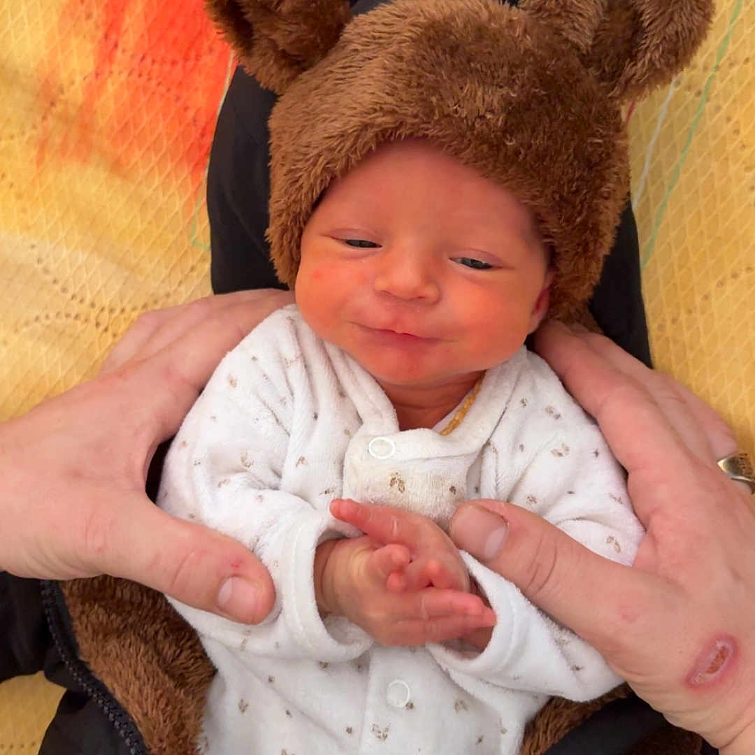 Maïwayne participe au concours pour gagner de l'argent avec cette photo : baby, infant, hands, onesie, hat, bear_ears, cute, newborn, sleepy, cozy, white_clothing, adult_hands, patterned_background, soft_texture, smiling, portrait, indoor, warm_colors, closeup, child