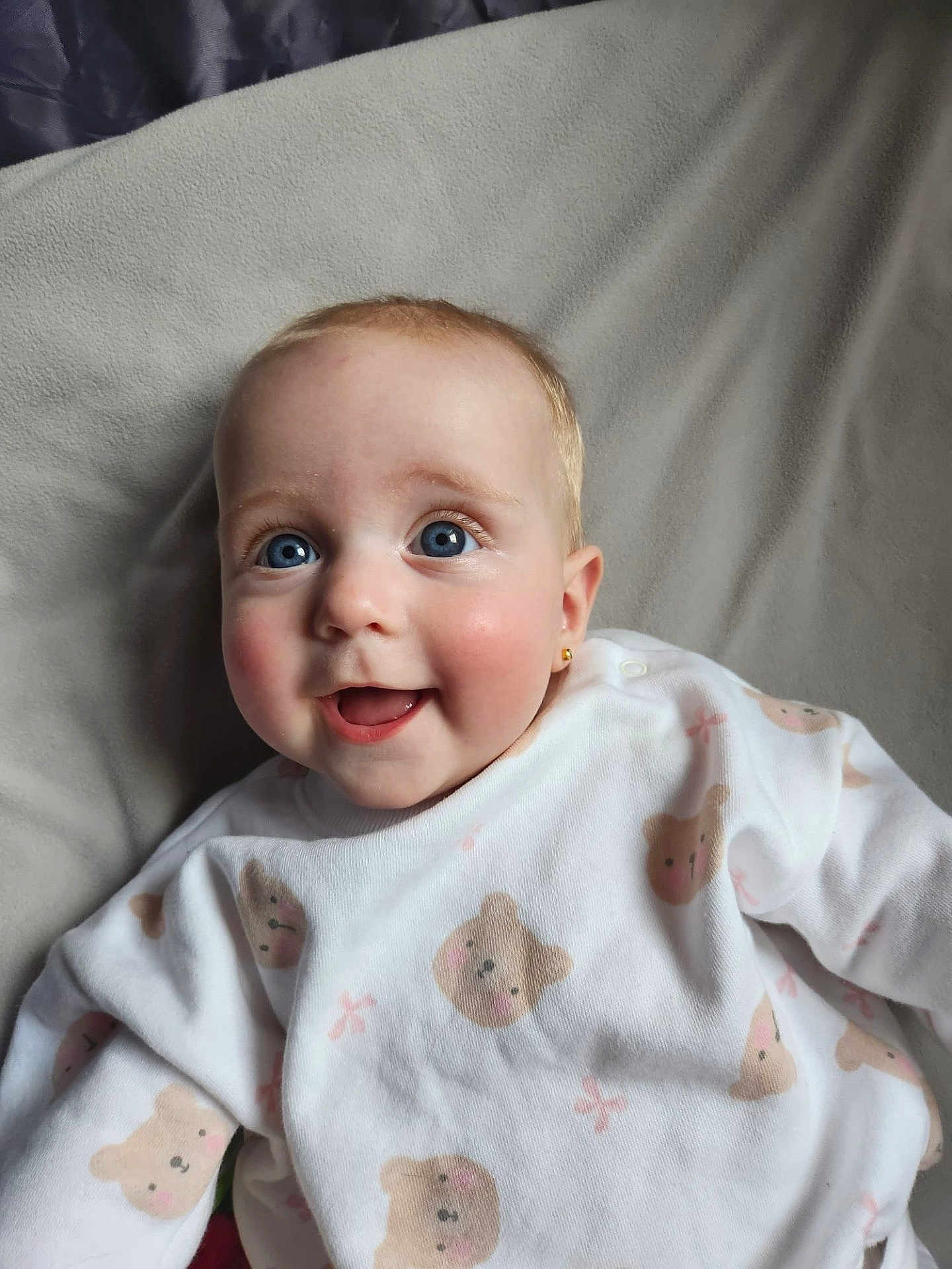 Leni-Rose is registered to the contest to win money with this photo: baby, blue_eyes, smiling, onesie, teddy_bear_pattern, cute, infant, lying_down, soft_blanket, white_clothing, happy, child, portrait, indoors, close_up, skin, face, earrings, cheeks, young