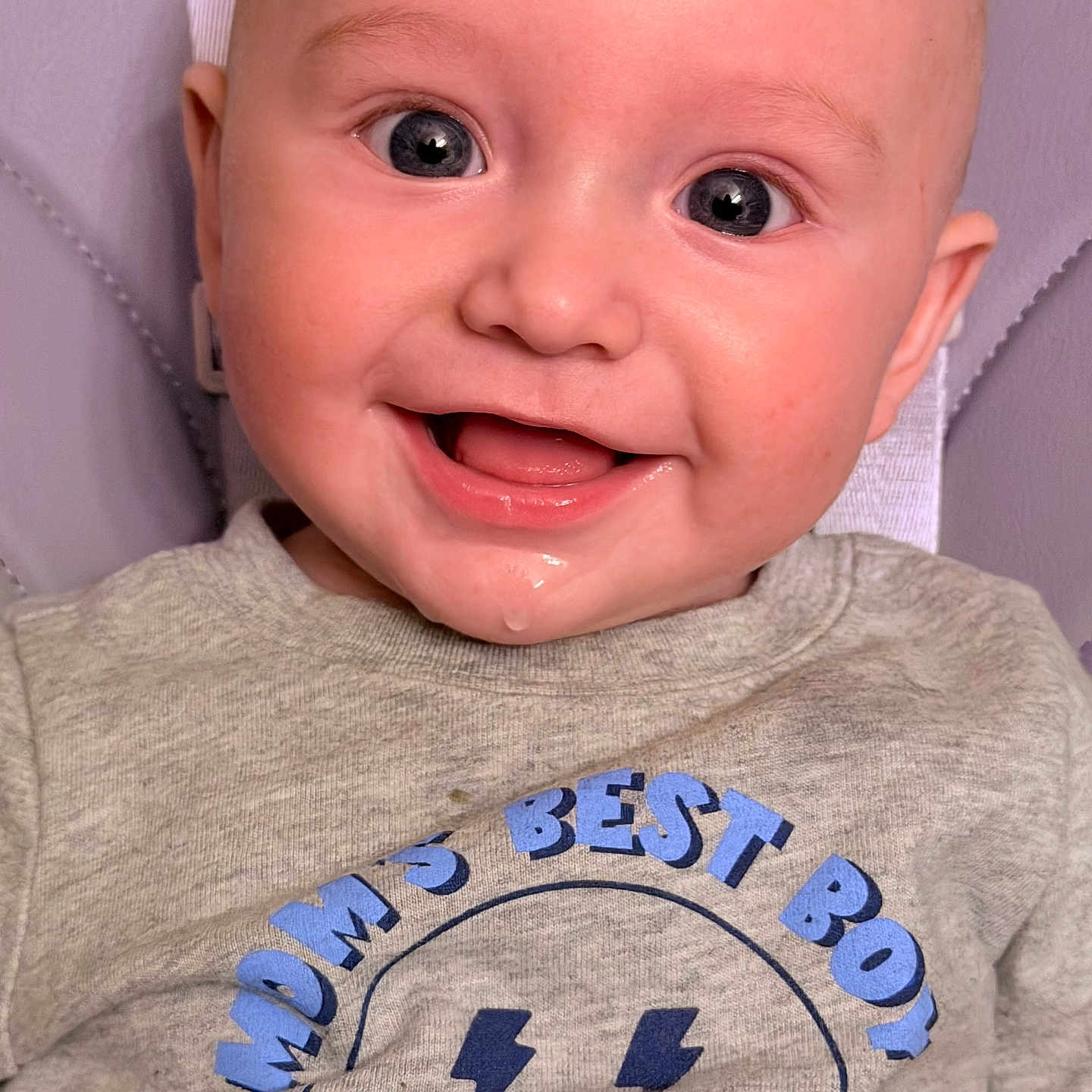 Rowan is registered to the contest to win money with this photo: baby, background, blue, child, closeup, clothing, cute, drool, eyes, face, gray, happy, head, infant, portrait, seated, shirt, skin, smile, text