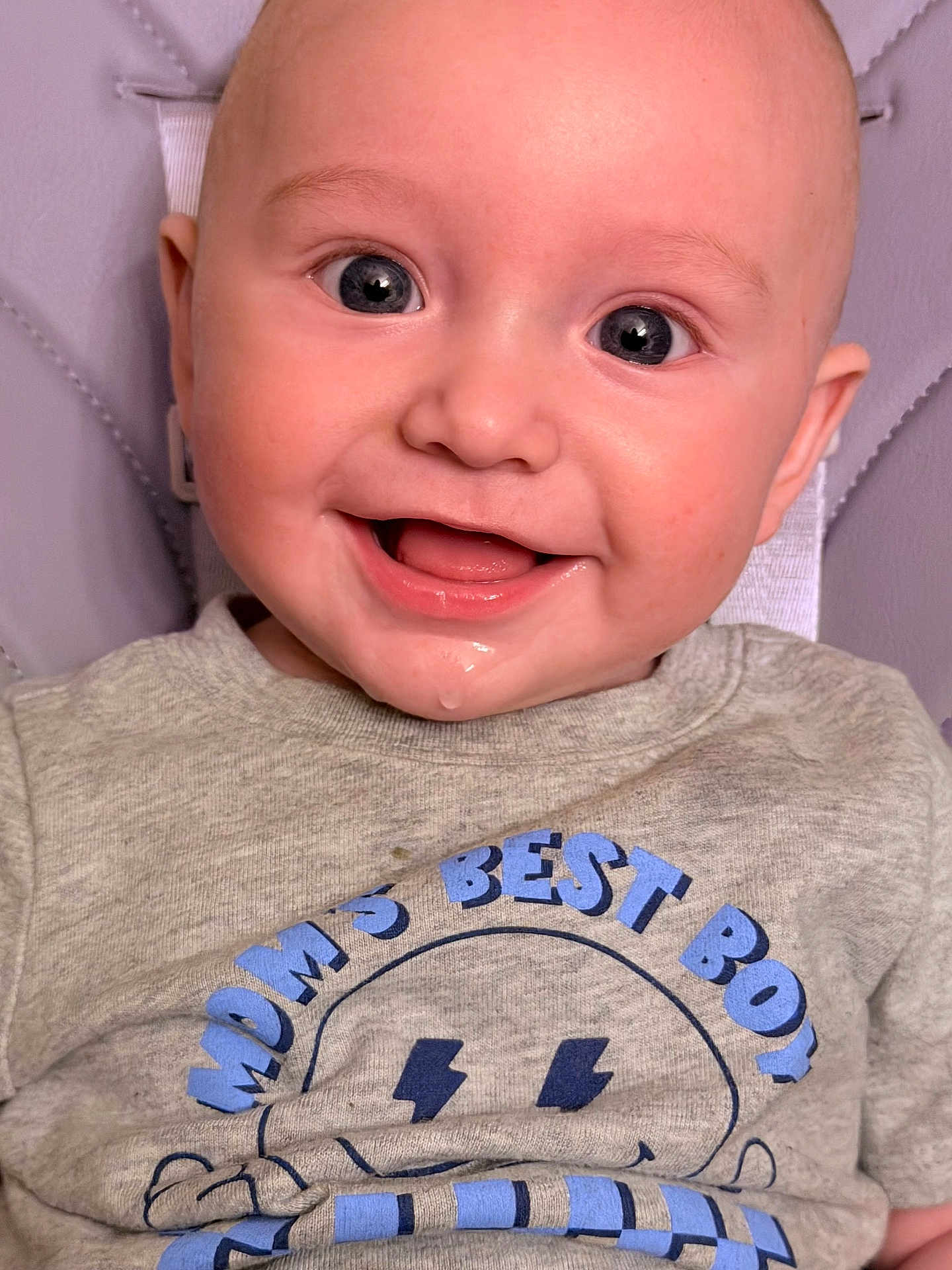 Rowan is registered to the contest to win money with this photo: baby, infant, smile, face, clothing, shirt, text, drool, happy, child, cute, portrait, seated, closeup, blue, gray, eyes, skin, head, background