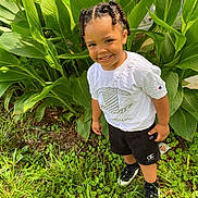 Kamarion is registered to the contest to win money with this photo: child, smiling, outdoor, greenery, plants, grass, white_shirt, black_shorts, braided_hair, young_child, happy, nature, casual_clothing, footwear, portrait, sunlight, daylight, standing, person, fashion