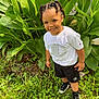 child, smiling, outdoor, greenery, plants, grass, white_shirt, black_shorts, braided_hair, young_child, happy, nature, casual_clothing, footwear, portrait, sunlight, daylight, standing, person, fashion