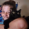 person, cat, black_cat, indoor, hug, affection, wide_eyes, human_face, pet, animal, smile, close_up, arm, hair, expression, feline, domestic, companionship, shoulder, background