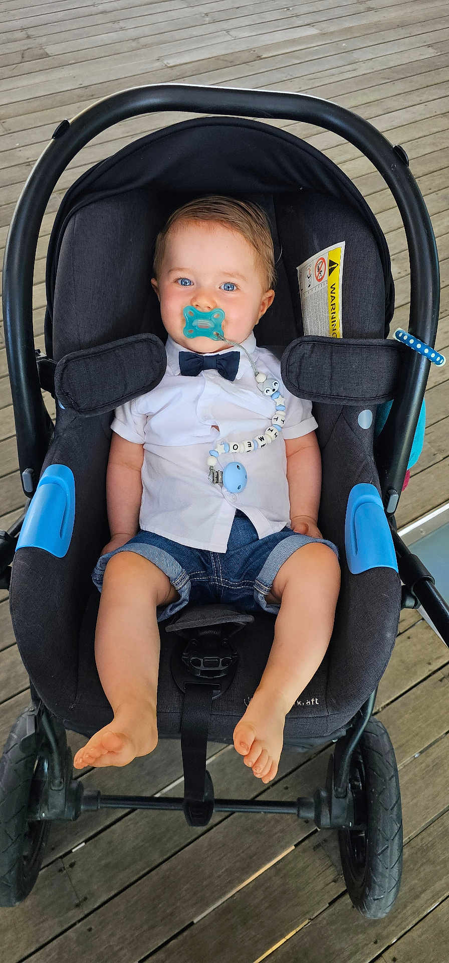 Timéo a rejoint le concours — aidez-le/la à gagner de superbes lots ! baby, child, stroller, pacifier, bow_tie, shirt, denim_shorts, beaded_clip, blue_eyes, sitting, wooden_deck, footwear, cute, infant, outdoor, baby_boy, clothing, face, portrait, baby_accessory