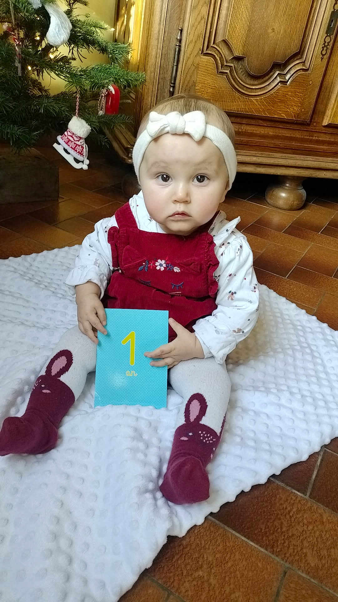 Elhya a rejoint le concours — aidez-le/la à gagner de superbes lots ! baby, blanket, bunny_socks, card, child, christmas_tree, cute, decor, floor, headband, holiday, indoor, number_one, ornament, portrait, red_clothing, sitting, soft_texture, tile, wooden_furniture