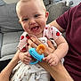baby, pacifier, adult, couch, blanket, clothing, face, hand, smile, tongue, infant, person, indoor, furniture, fabric, holding, pattern, happy, seated, skin