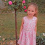 child, girl, pink_dress, floral_dress, rose_bush, flowers, garden, grass, smile, standing, portrait, outdoor, young_child, shoes, sneakers, foliage, tree_stem, summer, leaf_litter, cute