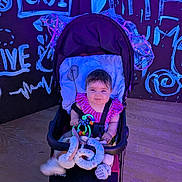 Laylah Mae is registered to the contest to win money with this photo: baby, stroller, toy, child, indoor, smiling, colorful, clothing, face, seat, floor, graffiti, background, wall, wooden_floor, person, happy, playful, infant, cute