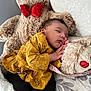 baby, sleeping, teddy_bear, stuffed_animal, yellow_clothing, red_bow, bed, blanket, pillow, animal_print, infant, child, cozy, cuddly, soft_toy, cute, peaceful, rest, indoors, portrait