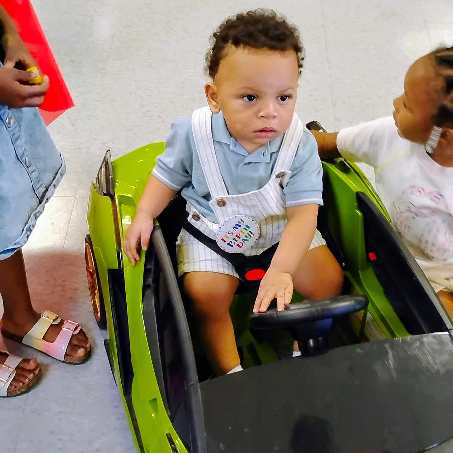 Ja'Karta joined the competition — help win amazing prizes! birthday_badge, child, clothing, curly_hair, earrings, floor, footwear, green, hair, hand, indoor, interaction, person, playing, seatbelt, shirt, shorts, steering_wheel, toddler, toy_car