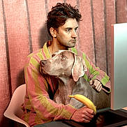 Paco participe au concours pour gagner de l'argent avec cette photo : man, dog, computer, desk, banana, bathrobe, chair, keyboard, mouse, digital_tablet, curtain, indoor, pet, technology, focused, seated, home_office, brown_dog, striped_clothing, looking