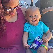 Romeo is registered to the contest to win money with this photo: baby, child, adult, glasses, smile, snack, blue_onesie, dinosaur_print, tattoo, bracelet, stroller, outdoor, sunlight, table, happy, person, holding, sitting, casual, family