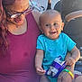baby, child, adult, glasses, smile, snack, blue_onesie, dinosaur_print, tattoo, bracelet, stroller, outdoor, sunlight, table, happy, person, holding, sitting, casual, family
