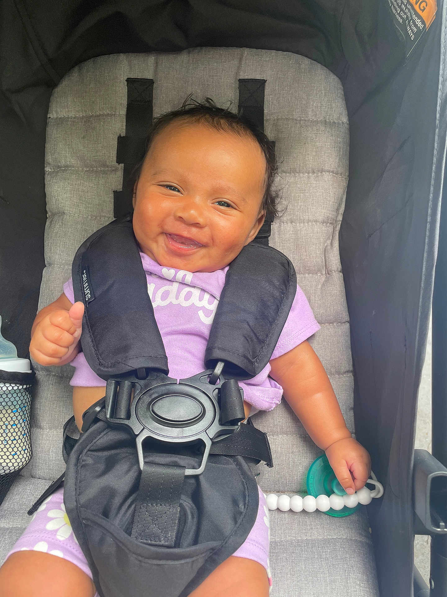 Deja is registered to the contest to win money with this photo: baby, smiling, stroller, harness, pacifier, purple_clothing, child, seat, happy, cute, infant, beads, safety, portrait, hand, face, comfort, outdoor, black, gray