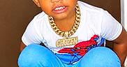 Chozen is registered to the contest to win money with this photo: child, toddler, boy, portrait, smiling, squatting, indoors, doorway, curly_hair, hairstyle, white_shirt, blue_pants, superhero_shirt, gold_chain, necklace, mismatched_socks, red_socks, black_mat, floor, fashion
