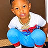child, toddler, boy, portrait, smiling, squatting, indoors, doorway, curly_hair, hairstyle, white_shirt, blue_pants, superhero_shirt, gold_chain, necklace, mismatched_socks, red_socks, black_mat, floor, fashion
