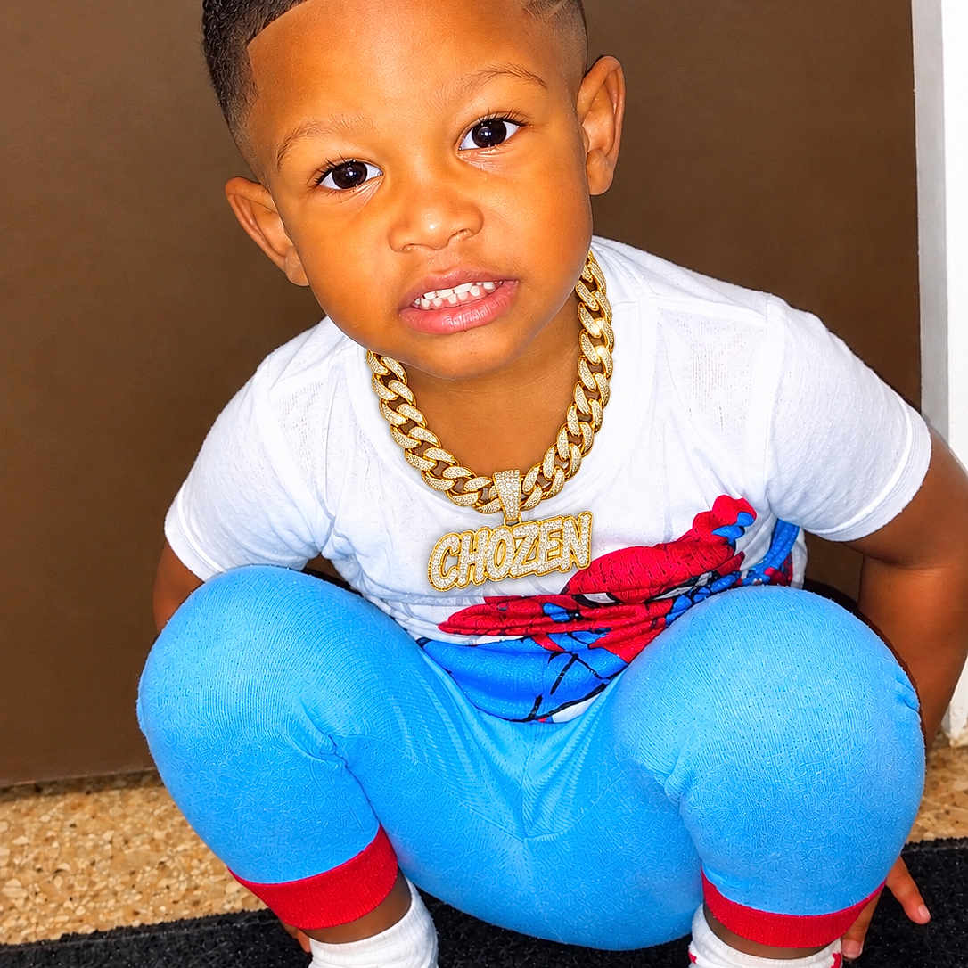 Chozen is registered to the contest to win money with this photo: child, toddler, boy, portrait, smiling, squatting, indoors, doorway, curly_hair, hairstyle, white_shirt, blue_pants, superhero_shirt, gold_chain, necklace, mismatched_socks, red_socks, black_mat, floor, fashion