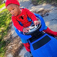 Chozen is registered to the contest to win money with this photo: child, toddler, spiderman, costume, red_suit, superhero, toy_car, steering_wheel, outdoor, park, sunlight, path, grass, trees, portrait, sitting, driving, playtime, helmet, phone_screenshot