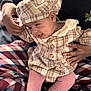 baby, beanie, bodypart, bonnet, cap, clothing, face, finger, hand, hat, head, newborn, pants, person, sitting, sunhat, tartan